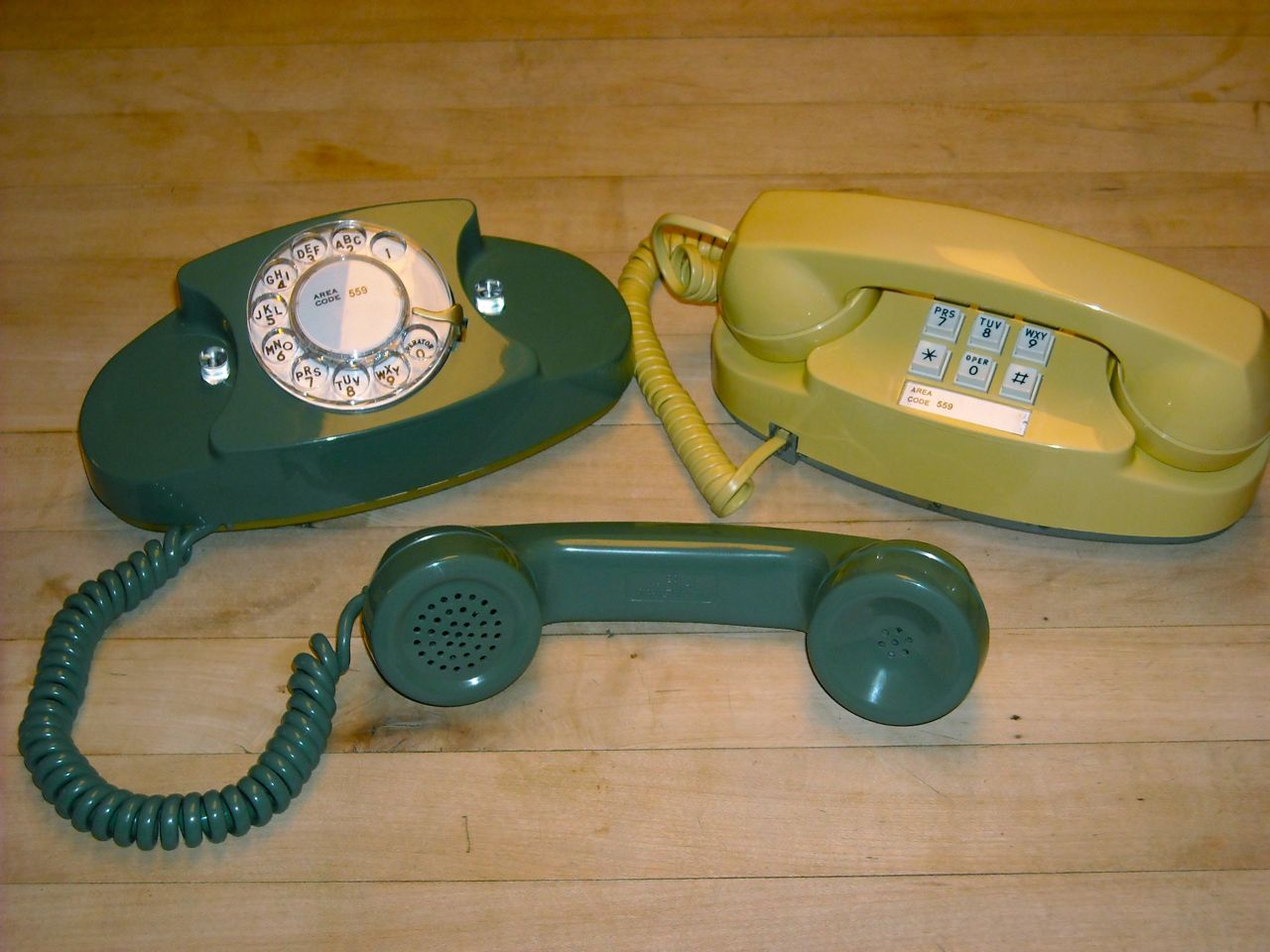 Two Princess telephones on a wooden table, a dark green rotary one with its handset off the hook and a pale yellow push-button one