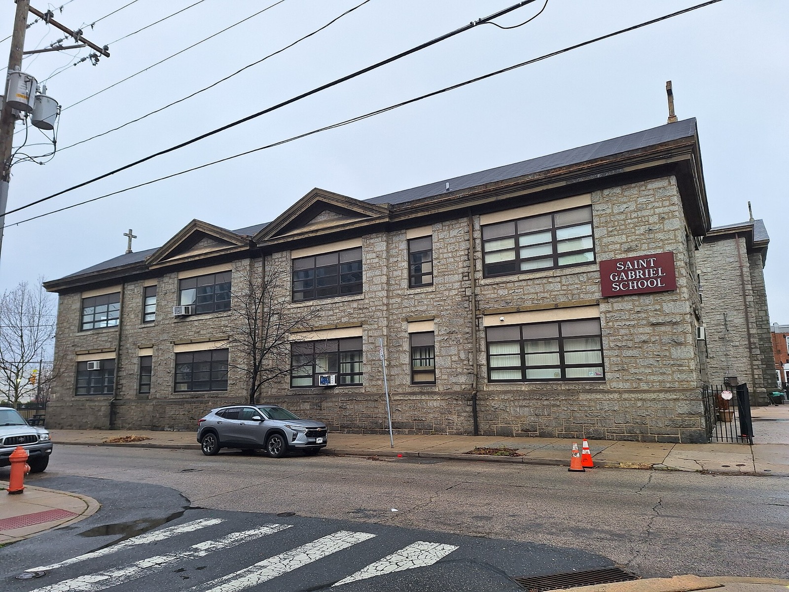 A two-storey grey stone school building on a street corner under overhead wires, with a maroon sign reading Saint Gabriel School on its end wall and a silver car parked outside