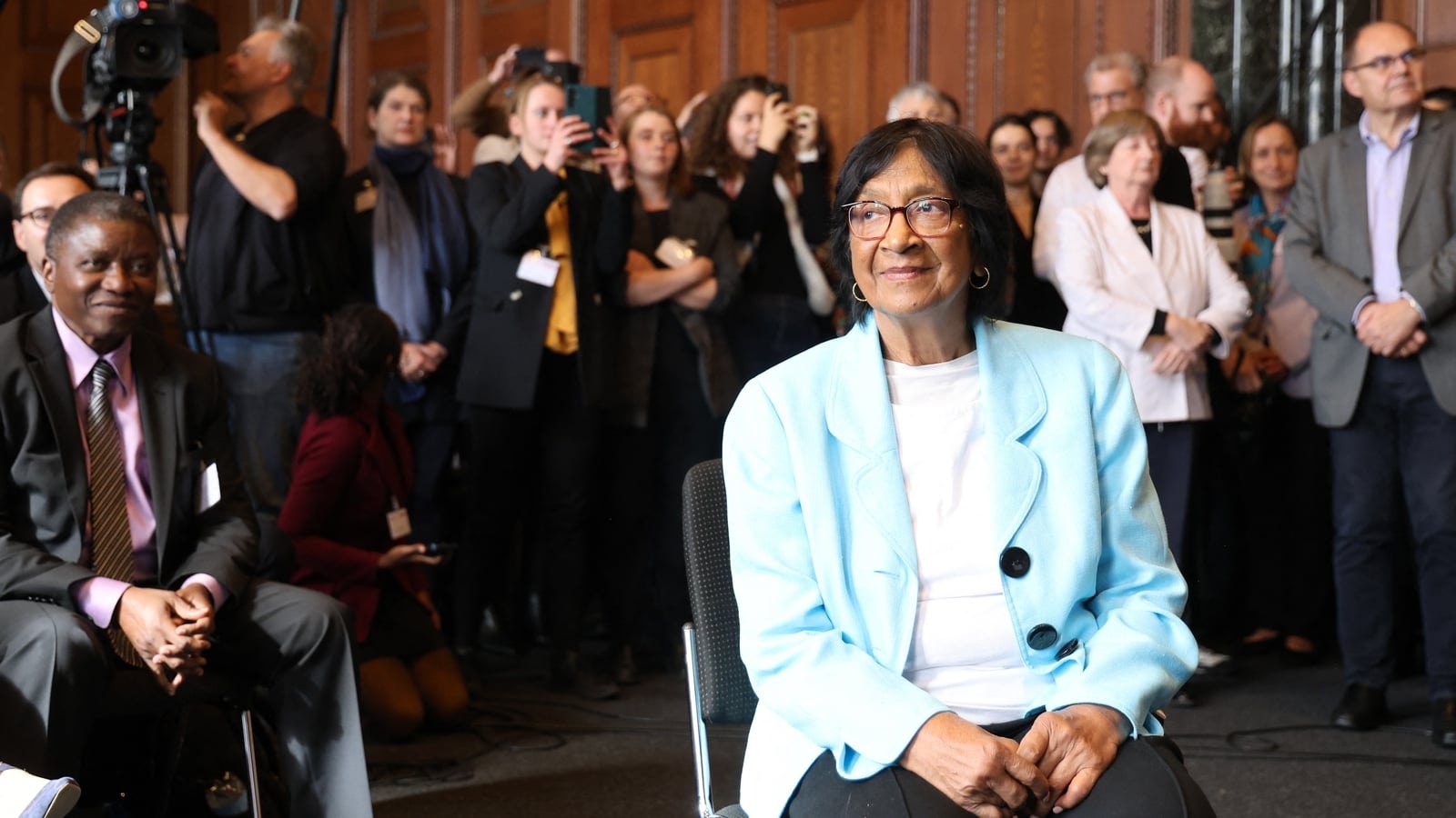 An older woman in a pale blue jacket and glasses sitting in a wood-panelled room, smiling, with a crowd of people and cameras standing behind her.