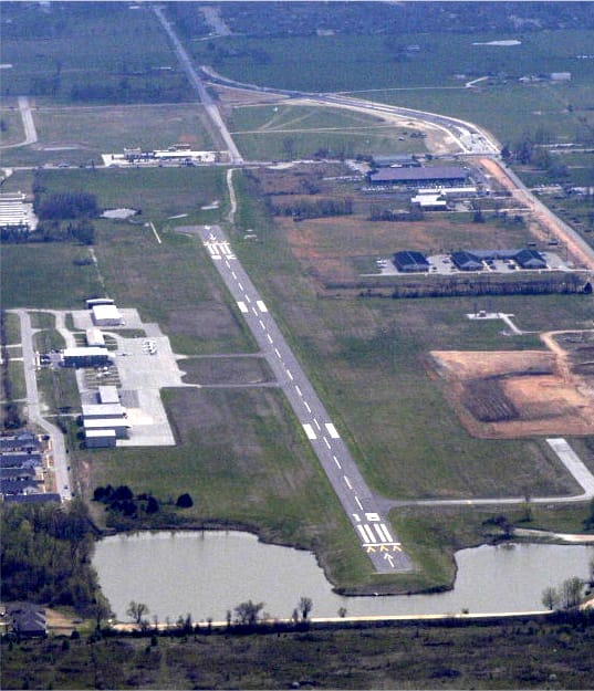 An aerial photograph of a single runway running diagonally between fields, with hangars on the left and a pond at the southern end