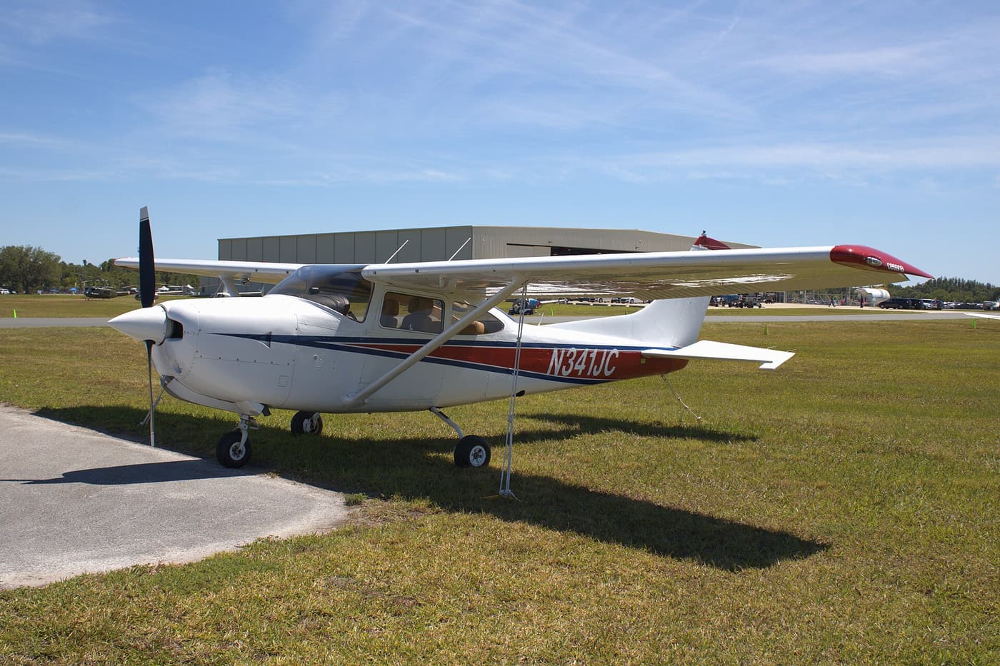 A white single-engine Cessna with red and blue stripes and the registration N341JC parked on grass, with its wing overhead and a hangar behind
