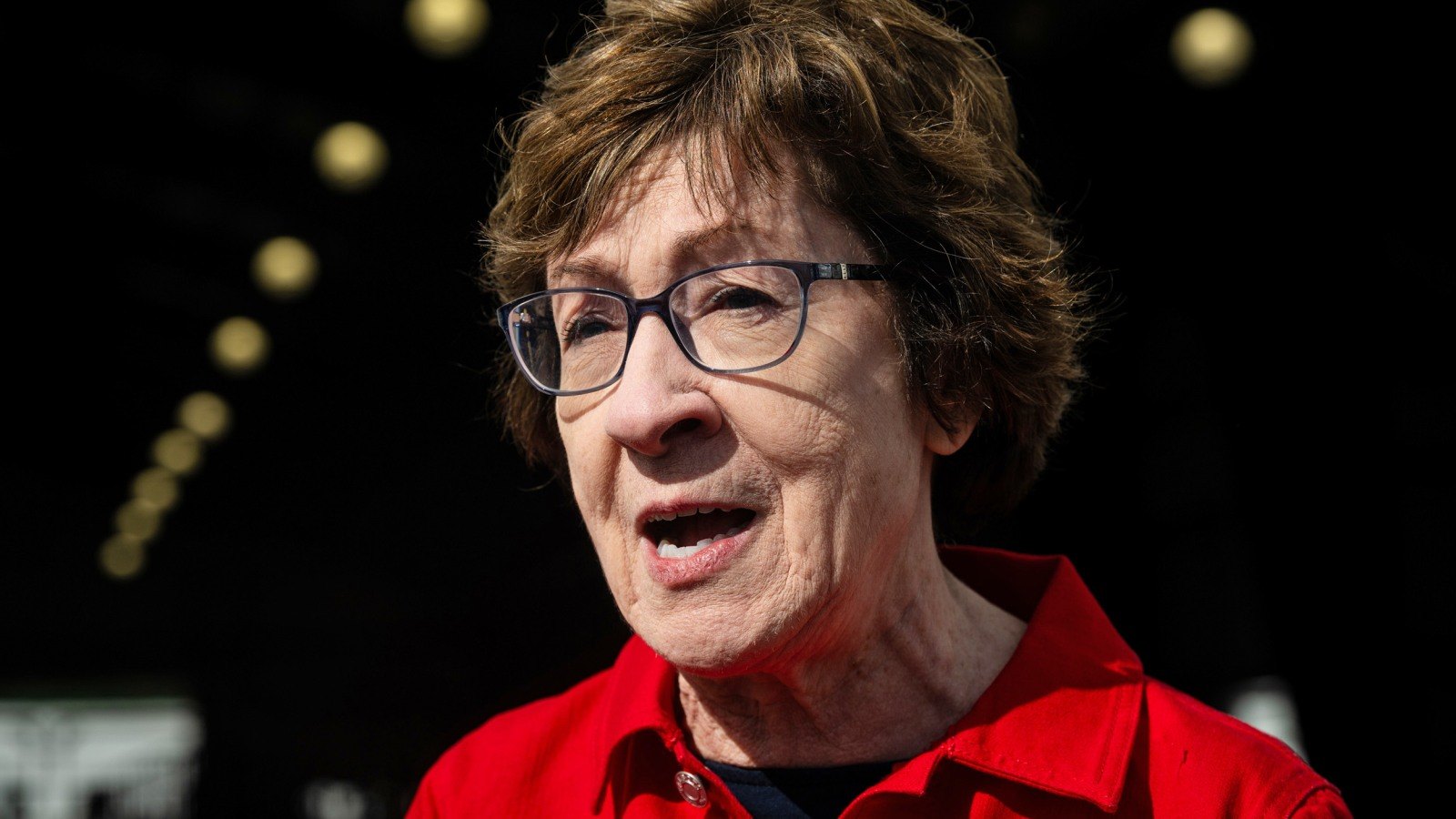 A woman with short auburn hair and dark-rimmed glasses, in a red jacket, speaking, with the lights of a barn roof blurred behind her.