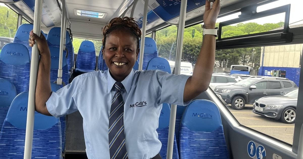 A smiling woman in a pale blue McGill's bus driver's shirt and striped tie, standing in the aisle of an empty bus and holding the grab rails on either side.