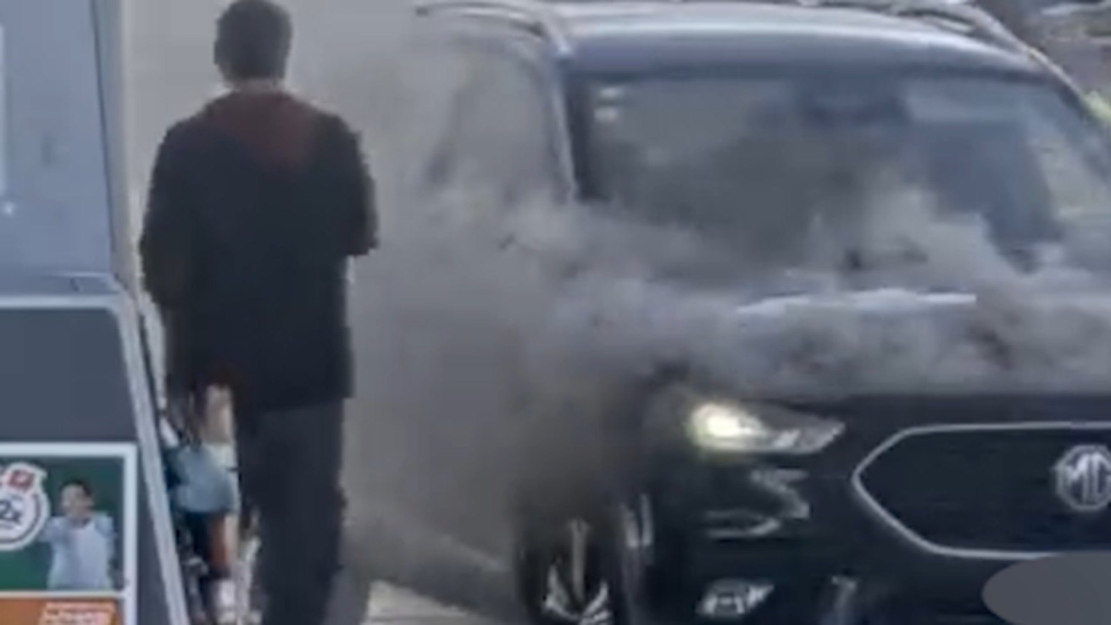 A grainy security-camera still: a man in a dark top walking toward a dark car at a petrol pump, with thick grey smoke pouring from the front of the car.