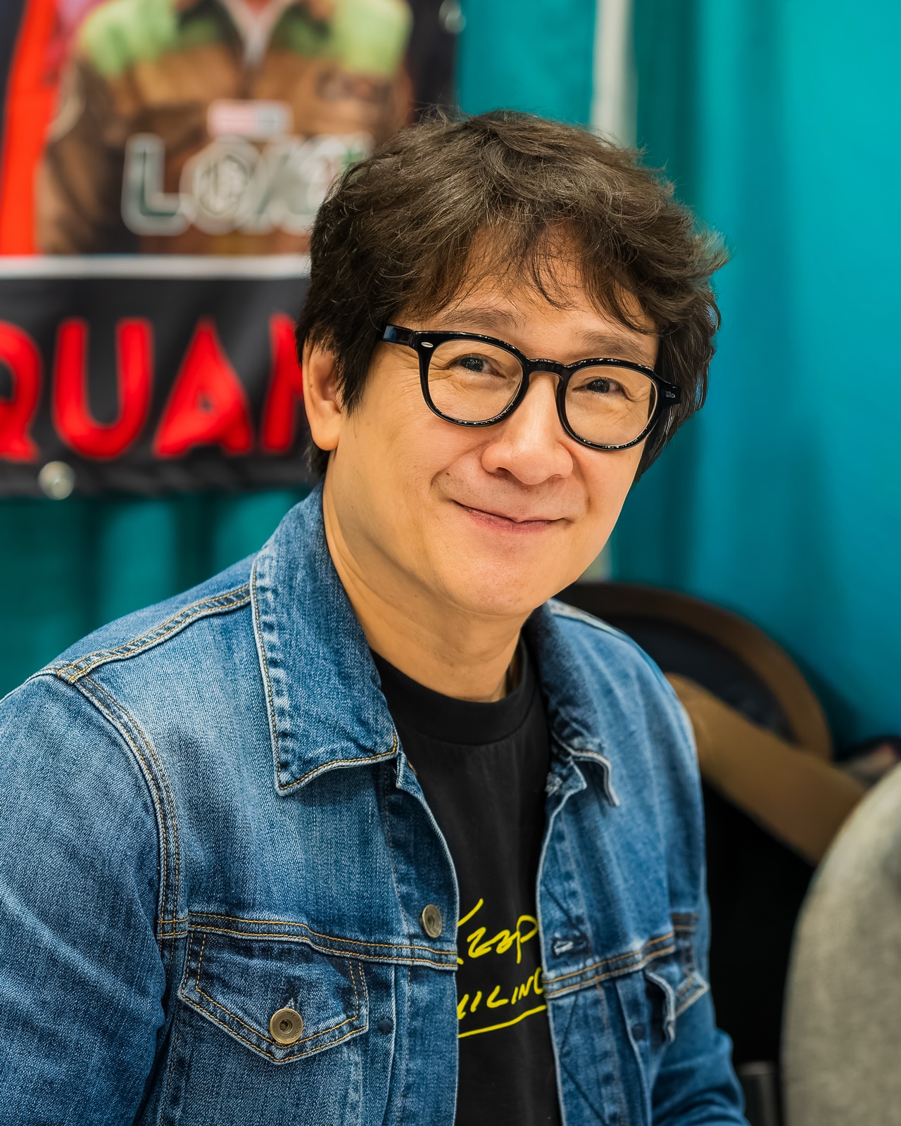 Ke Huy Quan sitting at a signing table in a denim jacket over a black t-shirt, wearing round black glasses and smiling at the camera, with a banner carrying his name on the wall behind him