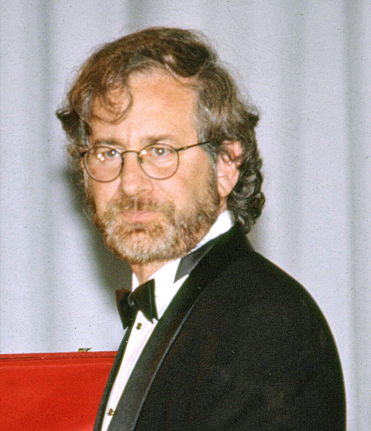 Steven Spielberg photographed in 1993 in a black dinner jacket and bow tie, with a beard and round wire-rimmed glasses, looking towards the camera