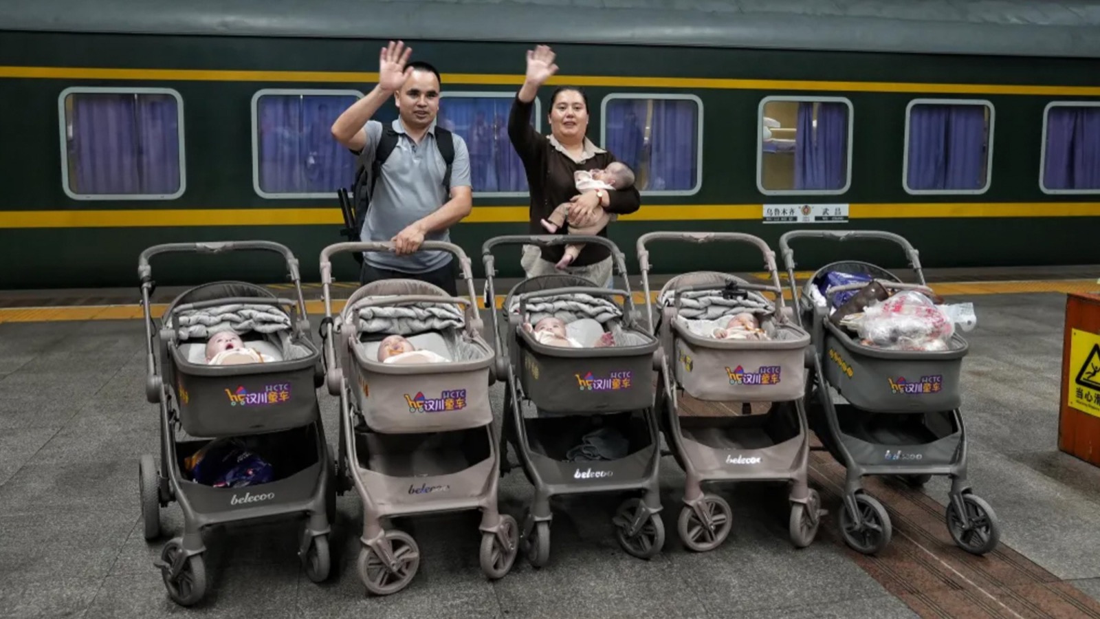 A man and a woman wave beside a green passenger train on a station platform. The woman holds a baby. In front of them stand five strollers, four of them holding sleeping babies.
