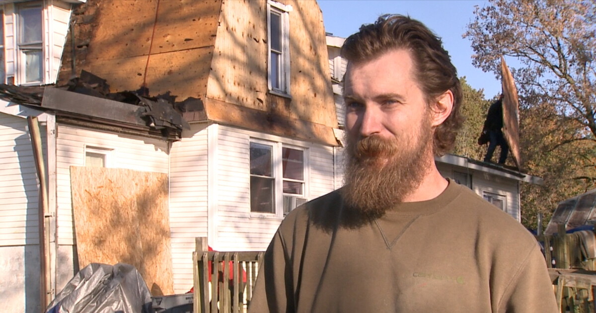 A bearded man in a brown sweatshirt stands in front of a white farmhouse whose roof has been stripped to bare wood, with a roofer working on top.