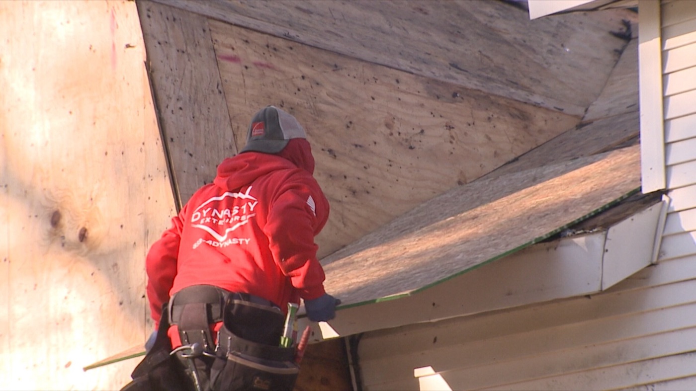 A roofer in a red Dynasty Exteriors hoodie and tool belt working on bare plywood on the farmhouse roof.