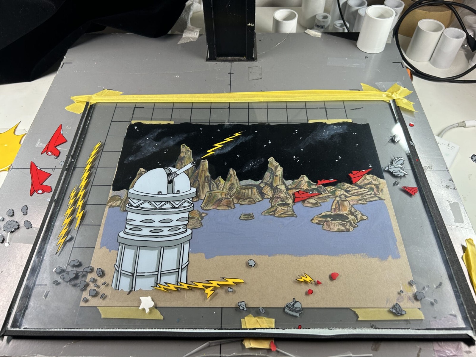 A hand-painted background of a rocky alien landscape with a white domed tower, laid flat on an animation table under a camera rig, with yellow tape, cut-out red ships and small painted rocks arranged on it