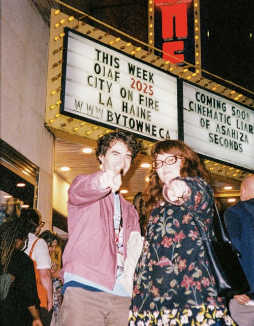 Brian Smee and Isabelle Aspin outside a cinema at night under a lit marquee, him in a jacket with a lanyard, her in a dark floral dress