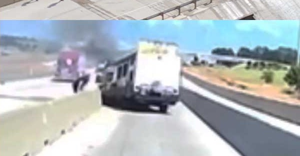 A frame from a lorry's dash cam looking along Interstate 30: a vehicle burning with pink and white flames against the central barrier, thick black smoke rising across the carriageway, a white camper trailer standing behind it and a figure on foot beside the camper