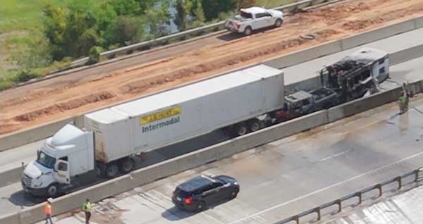 An aerial view of a closed Interstate 30: a white Intermodal semi-trailer stopped in the carriageway, and behind it against the concrete central barrier the burnt-out frame of a pickup and its trailer, with workers in high-visibility jackets standing on the empty road