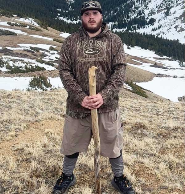 A young bearded man in a camouflage hoodie and khaki shorts stands on a bare hillside with patches of snow behind him, holding a long wooden walking stick in both hands
