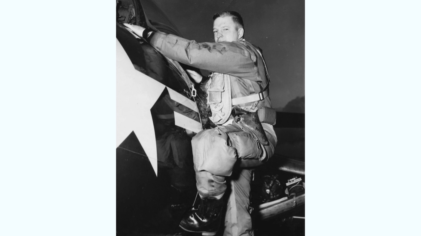 A black-and-white photo of a young pilot in flight gear climbing into the cockpit of a Navy jet marked with a white star.