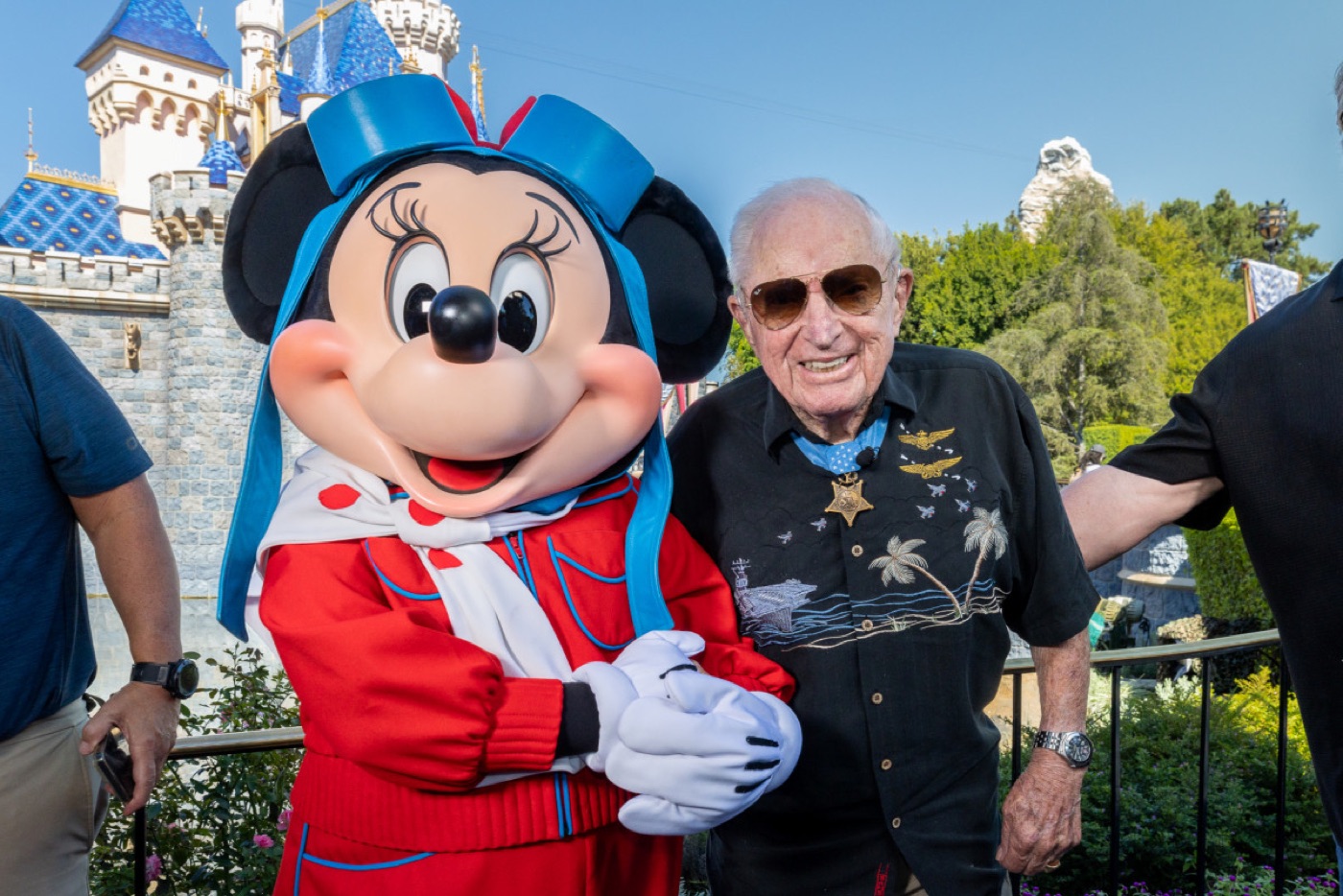 An elderly man in sunglasses and a black shirt with a medal around his neck stands arm in arm with Minnie Mouse in front of Sleeping Beauty Castle.