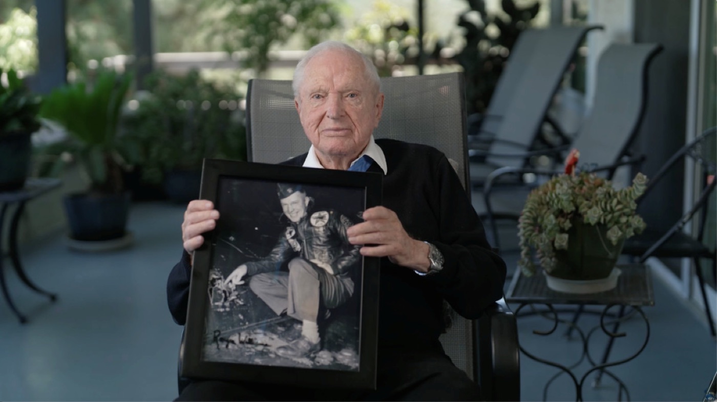 An elderly man in a black sweater sits on a sunny porch holding a framed black-and-white photo of himself as a young pilot climbing into a jet.