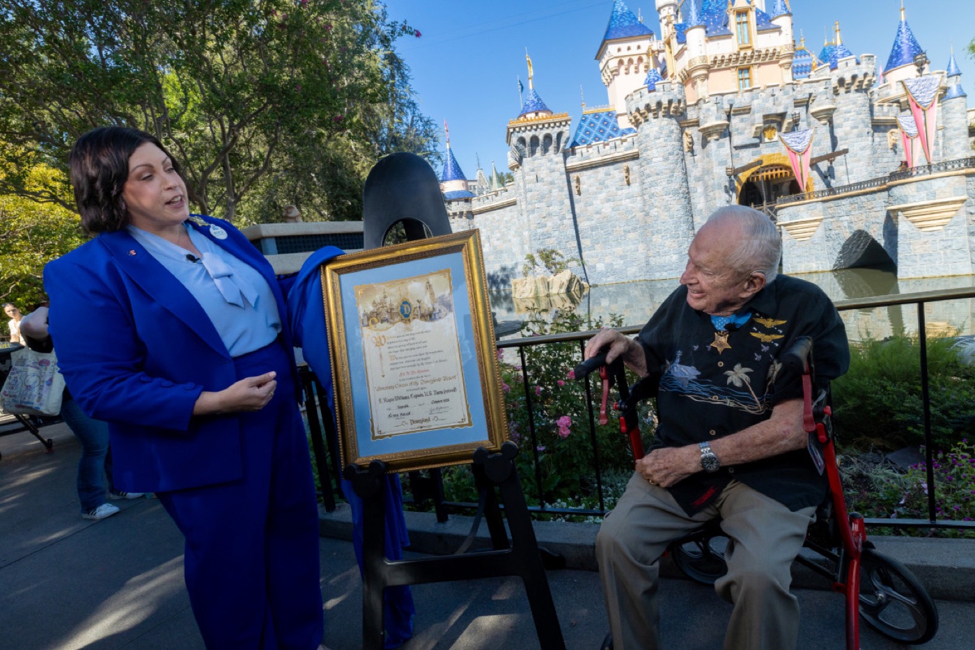 A woman in a blue suit stands beside a framed certificate on an easel, facing an elderly man with a walker, in front of a fairytale castle.