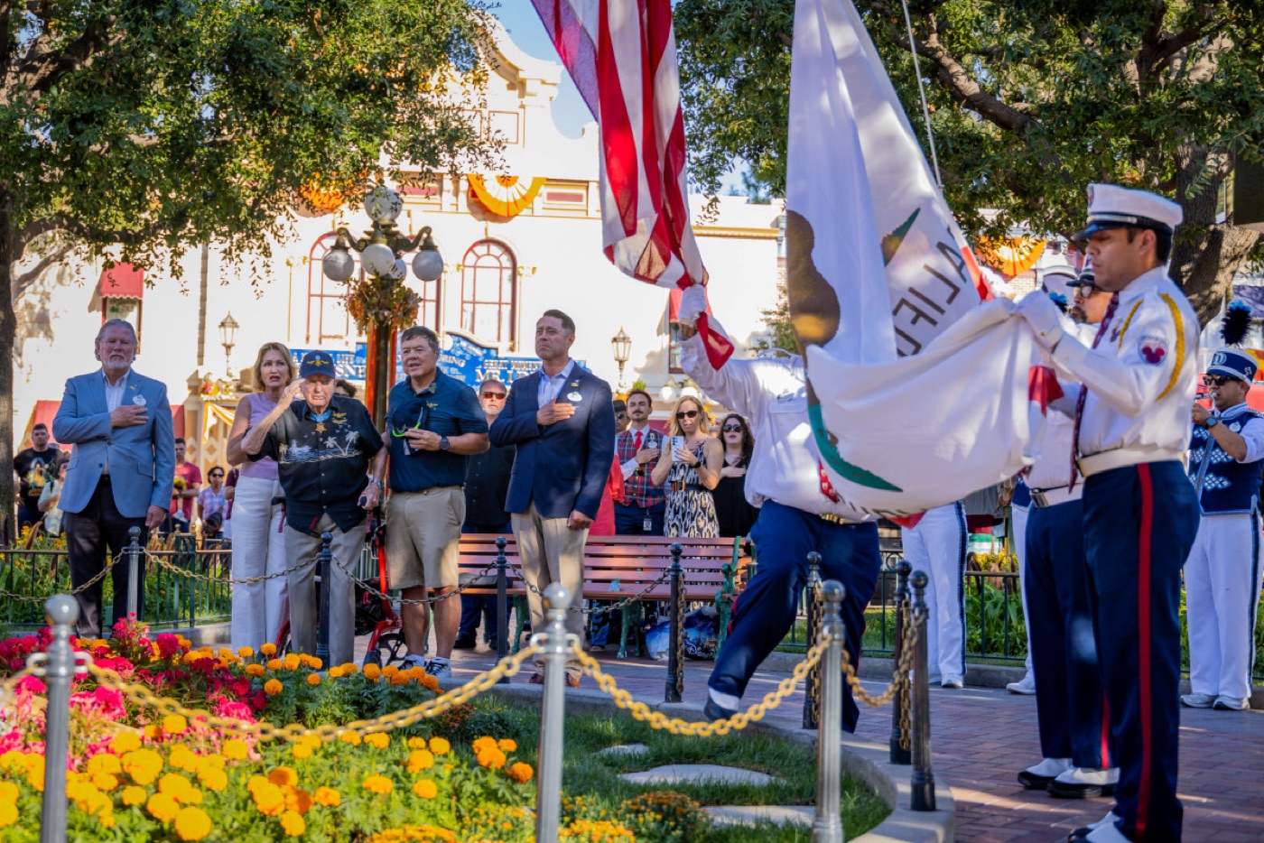 A colour guard lowers the American flag in a town square as a crowd stands with hands on hearts; an elderly man in a Navy cap stands among them.