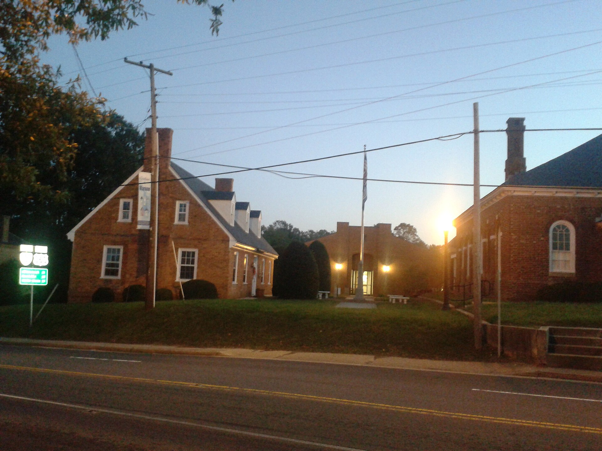 A small brick county courthouse complex in rural Virginia at dusk, with a flagpole, a street light coming on, overhead power lines and an empty road in the foreground