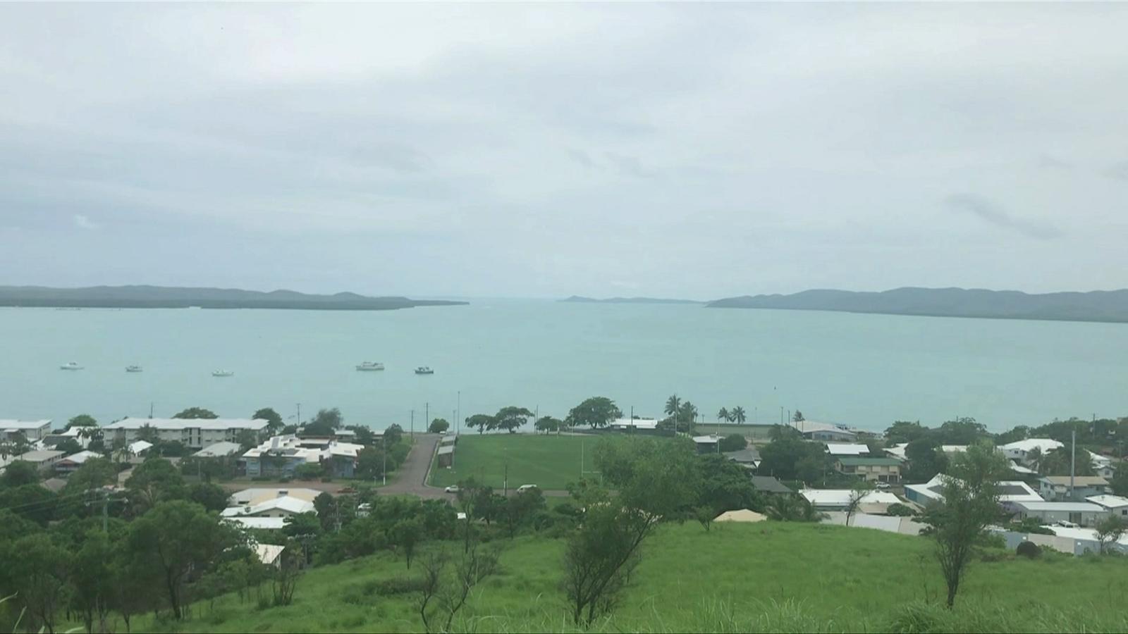 Thursday Island from a hill: tin roofs, a green oval by the water, boats at anchor on a flat grey-blue sea, and low islands on the horizon