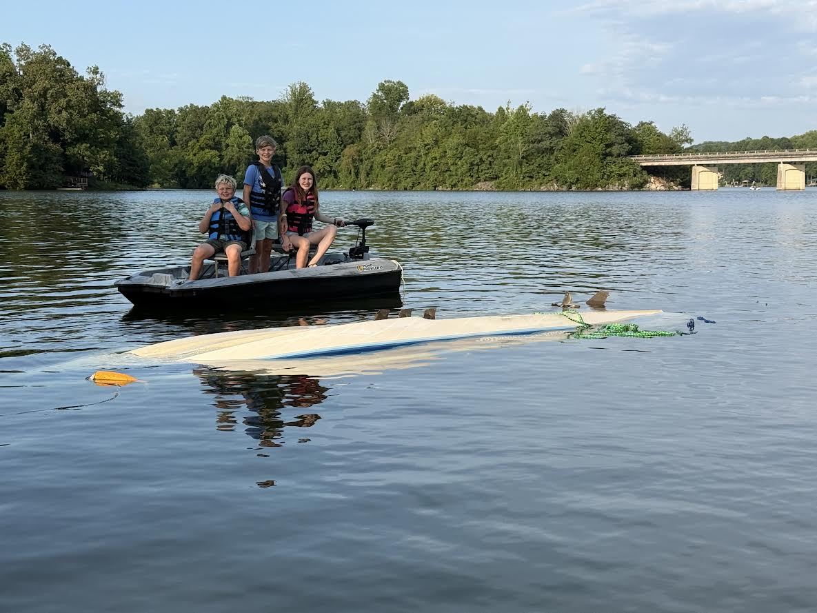 Three children in life jackets on a small flat fishing boat on a lake, beside an overturned white boat with only its hull above the water.