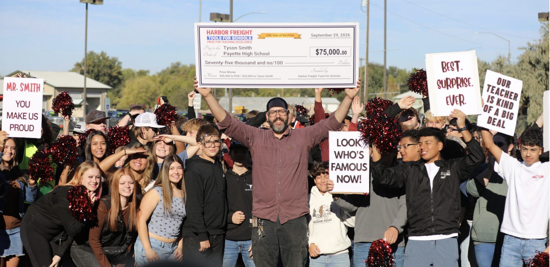 Tyson Smith, in a maroon shirt and glasses, holds a large check for $75,000 over his head. Students crowd around him with red and black pom-poms and handmade signs.