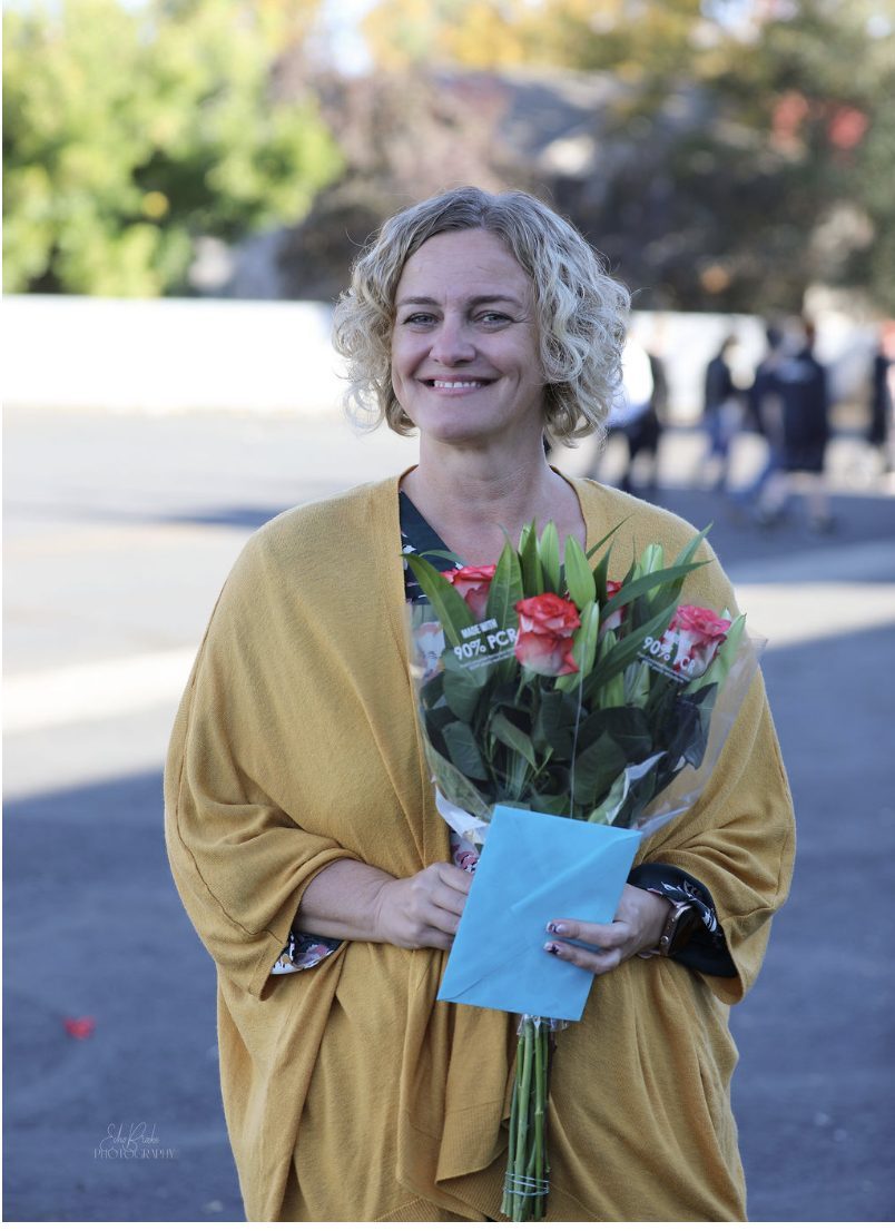 Amanda Smith, with short curly blonde hair and a mustard cardigan, smiles and holds a bunch of red roses and a light blue envelope.
