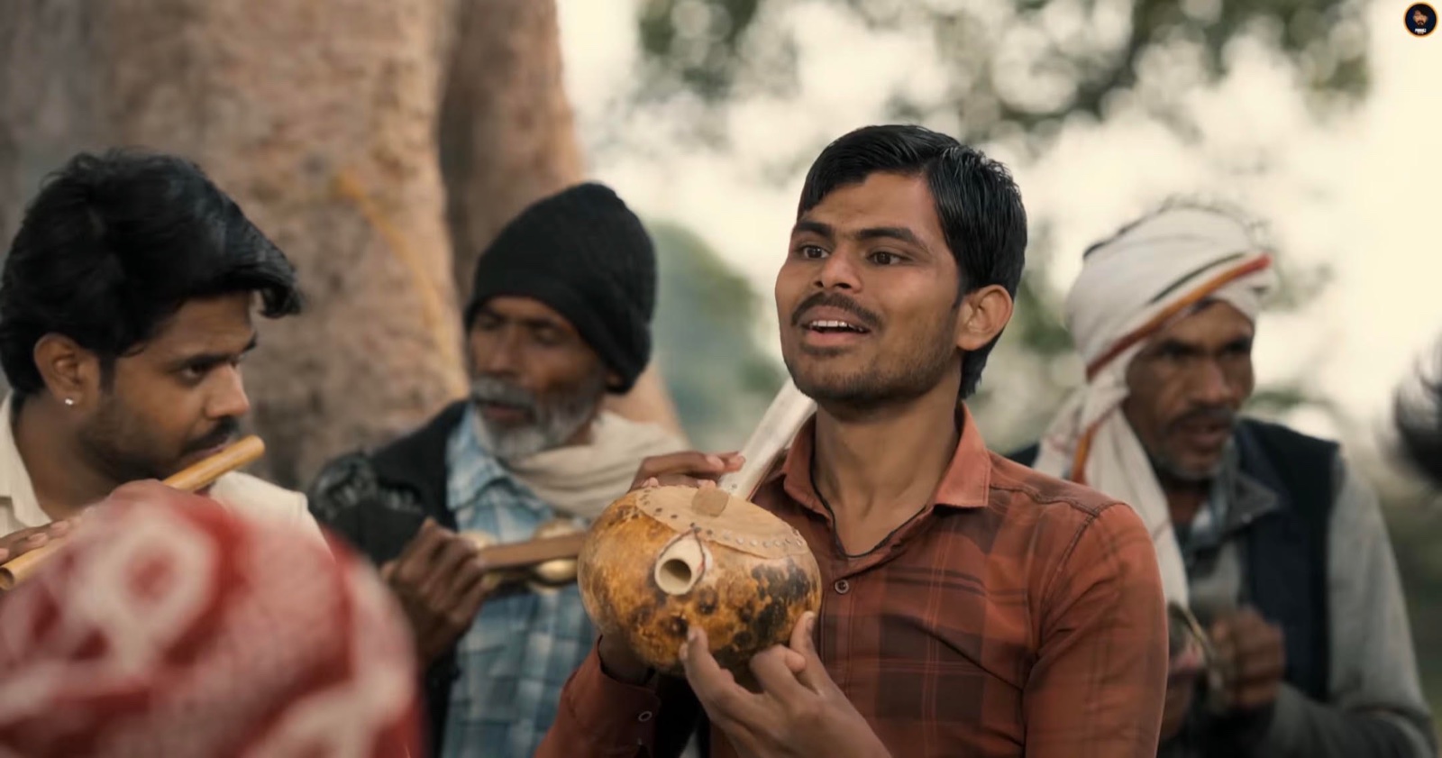Suneel Lodhi sings and plays his gourd-bodied tambura under a tree, with a flute player and older men in turbans around him