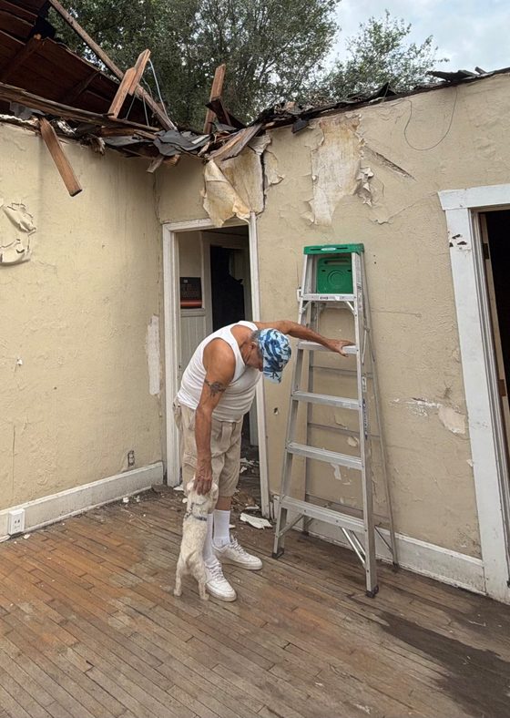 Gilbert Kirk, in a white vest and a blue cap, bending down to his small white dog on the bare wooden floor of a damaged room, a green stepladder against the wall beside him