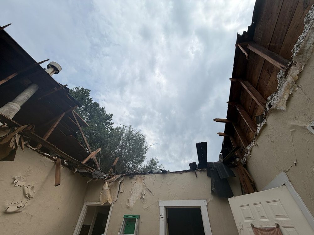 Looking up from inside the house: the ceiling and roof are torn away, broken rafters and boards hang at the edges, and grey sky fills the middle of the frame