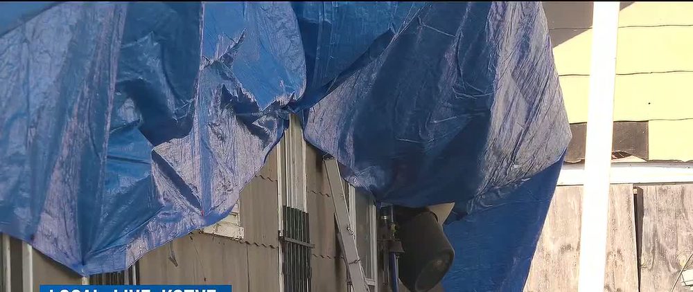 A new blue tarp stretched over the damaged side of the house, hanging down over a window and the wooden siding