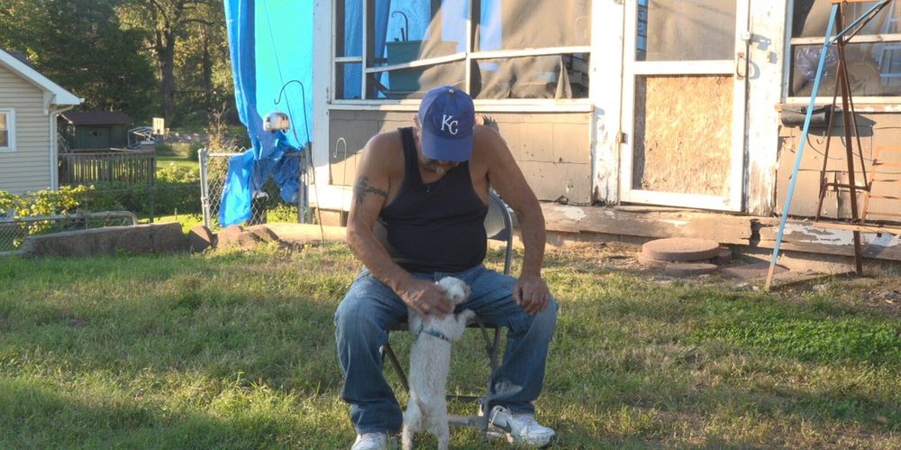 Gilbert Kirk, an older man in a blue Kansas City Royals cap and a black vest, sitting on a folding chair in his front yard and stroking his small white dog, with a blue tarp hanging over the side of his house behind them