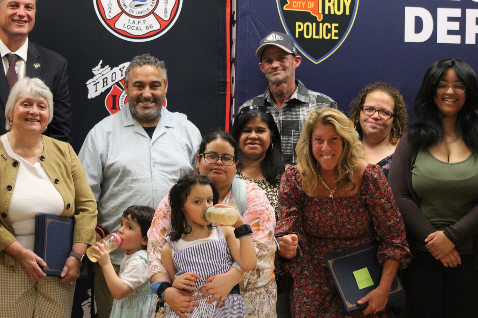 A group photograph at Troy city hall: the mayor and officials holding blue certificate folders, a family including several children with drinks bottles, and a man in a black and white check shirt and baseball cap standing at the back in front of a Troy Police Department banner