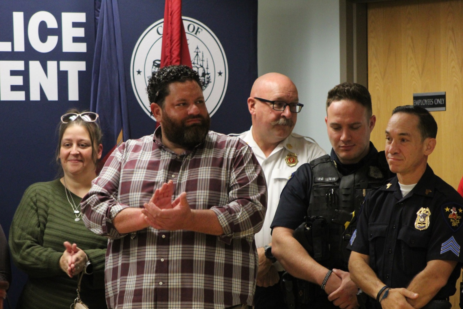 Michael Schnoop applauding in a plaid shirt at the ceremony, with a fire officer in a white shirt behind him and two Troy police officers in uniform to his right
