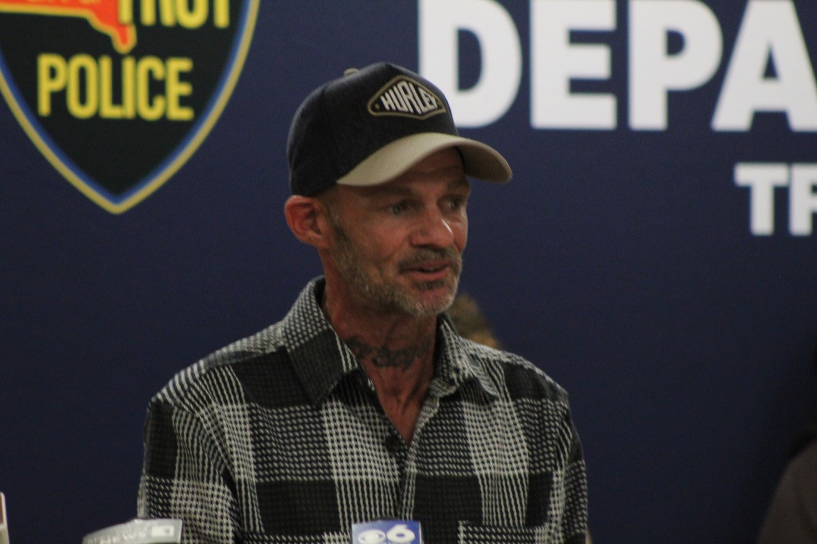 Michael Schnoop in a black and white check shirt and a dark baseball cap, speaking in front of a Troy Police Department banner with a television microphone in front of him