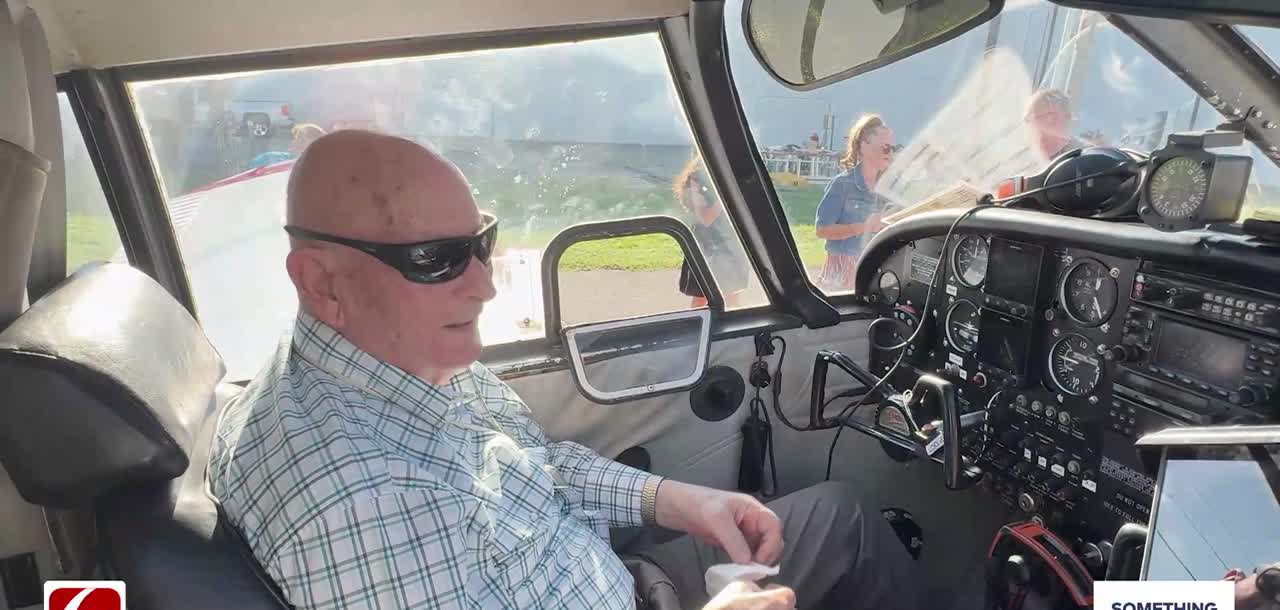 A bald man in his nineties in sunglasses and a checked shirt sits in the left seat of a small plane, the instrument panel beside him, with people visible through the cockpit window on the tarmac outside