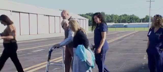 An elderly man pushes a walker across an airport apron with a woman at his side and nurses in blue scrubs walking alongside, hangars behind