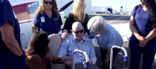 An elderly man in a chair beside a small plane, surrounded by women in hospice uniforms and family members with heads bowed and hands on his shoulders