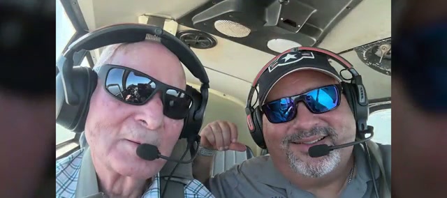 Two men in aviation headsets and sunglasses in the cockpit of a small plane in flight, the older man in a checked shirt on the left, the pilot in a grey polo on the right