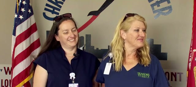 Two women in dark blue hospice uniforms with name badges stand in front of an American flag and a wall mural, one dark-haired, one blonde, both smiling