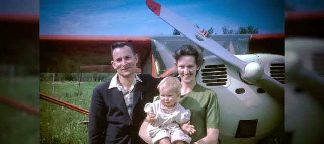 An old colour photograph of a young man in a dark jacket and a woman holding a baby, standing beside the nose and propeller of a small red-and-white plane on grass