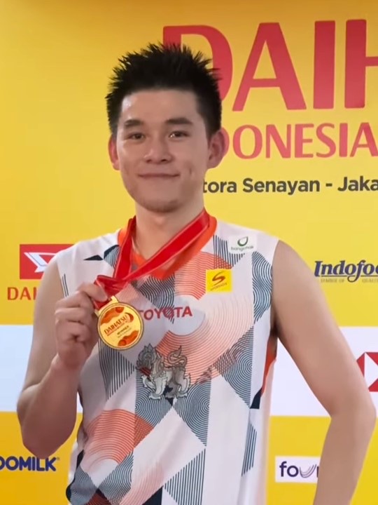Kunlavut Vitidsarn smiles and holds up a gold medal in front of a yellow sponsor board