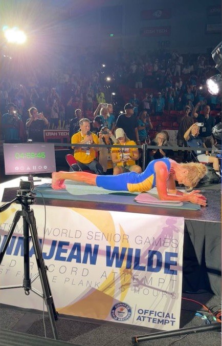The plank seen from the side of the stage: the timer at 04:59:45, photographers in yellow shirts in front of a packed arena, and a banner reading DonnaJean Wilde, record attempt, with the Guinness World Records logo.