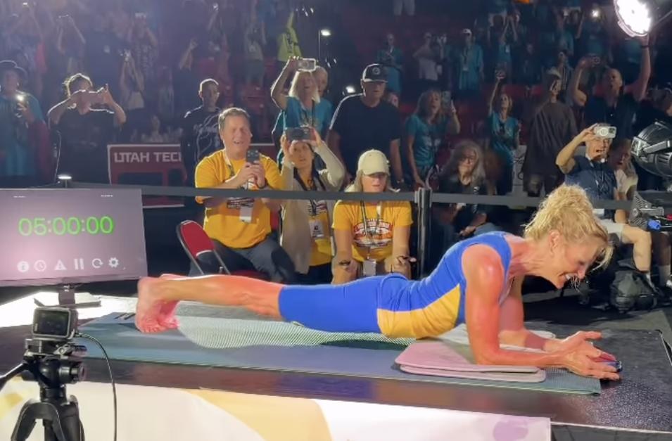 A woman in a blue and yellow suit holding a plank on her forearms on a raised stage, smiling, beside a timer reading 05:00:00, with a crowd behind her filming on their phones.