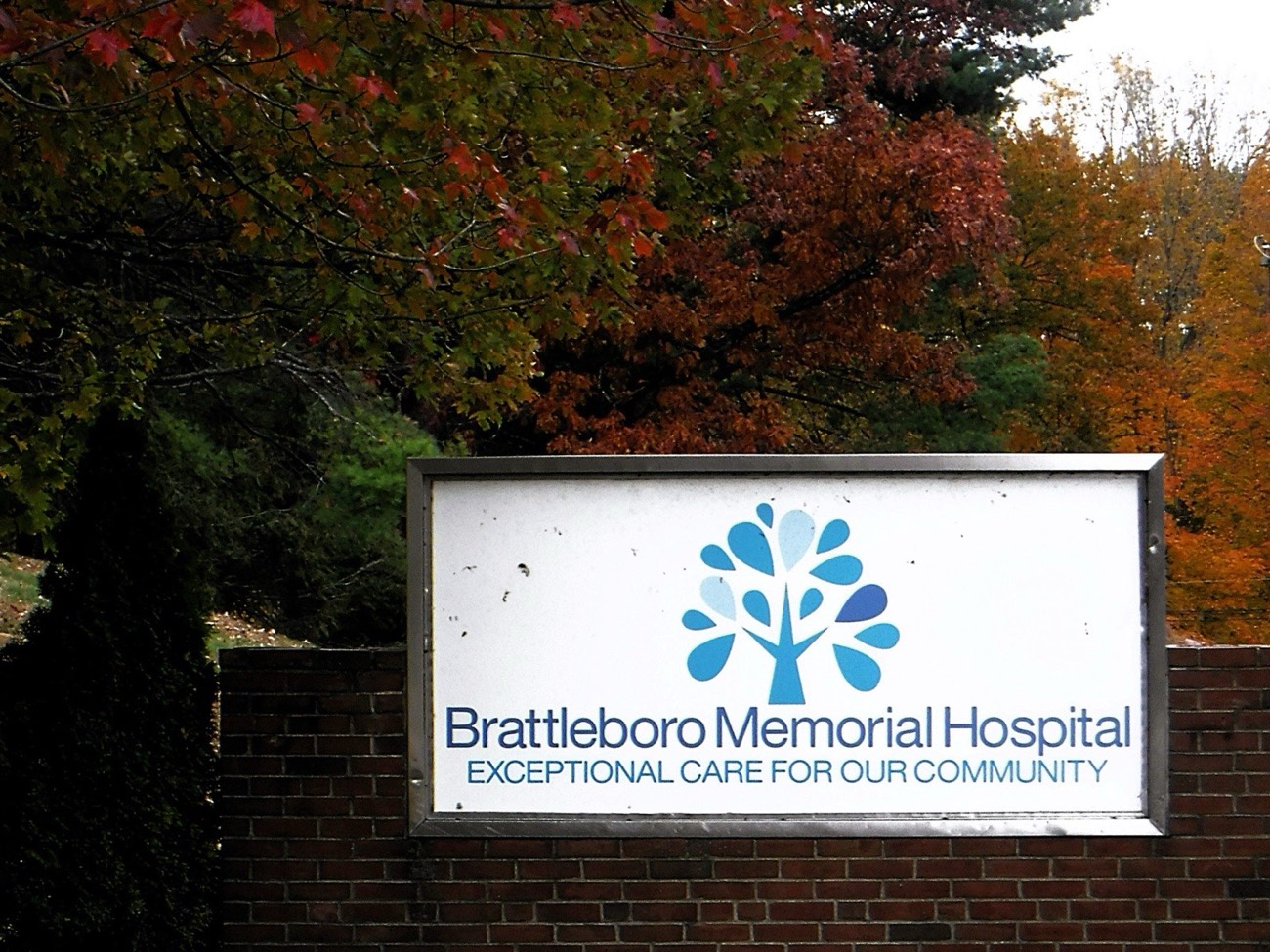 The Brattleboro Memorial Hospital sign on a brick wall, under trees in autumn colours.