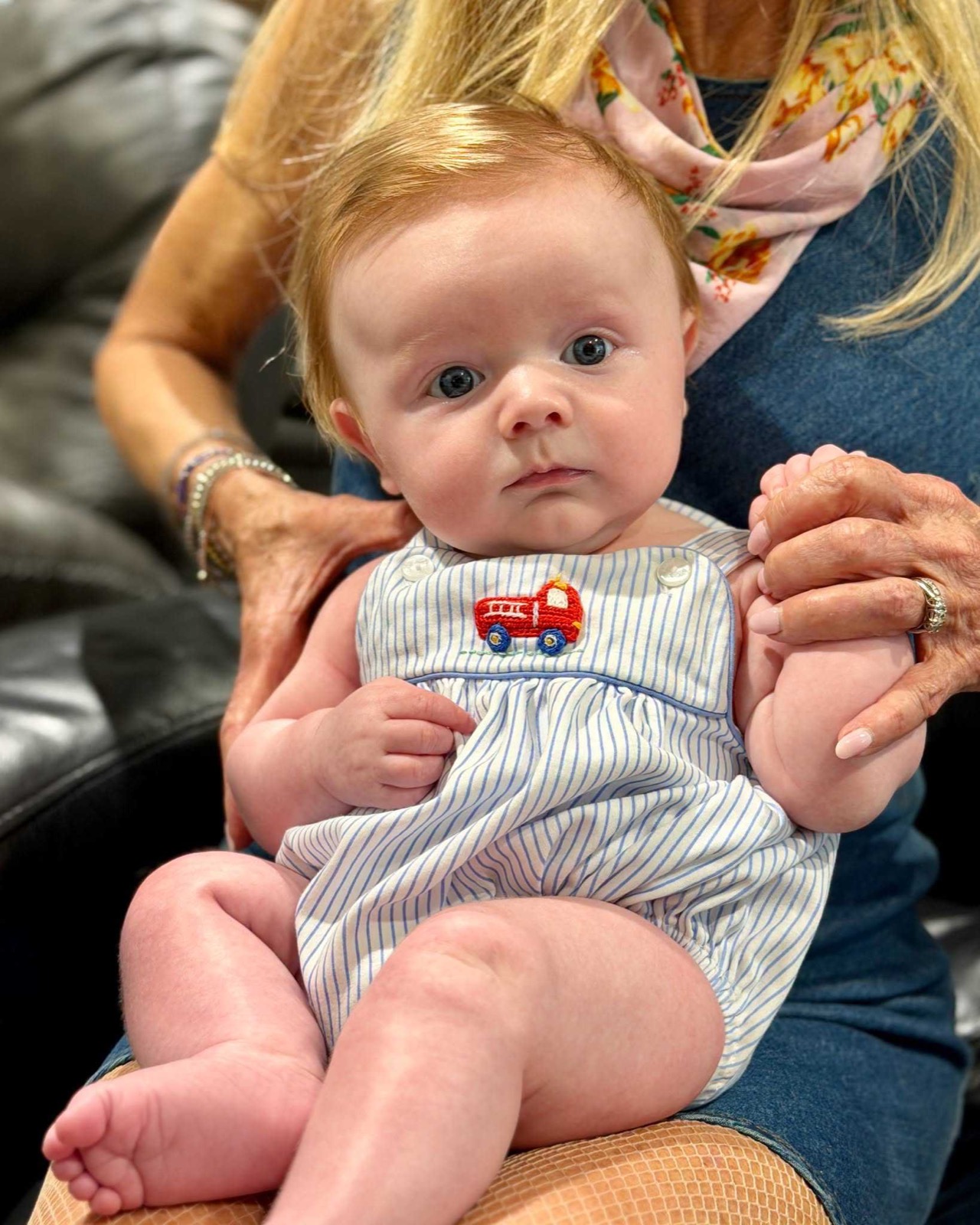 A red-haired baby with wide blue eyes sits on a woman's lap in a striped romper with a red fire engine embroidered on the front