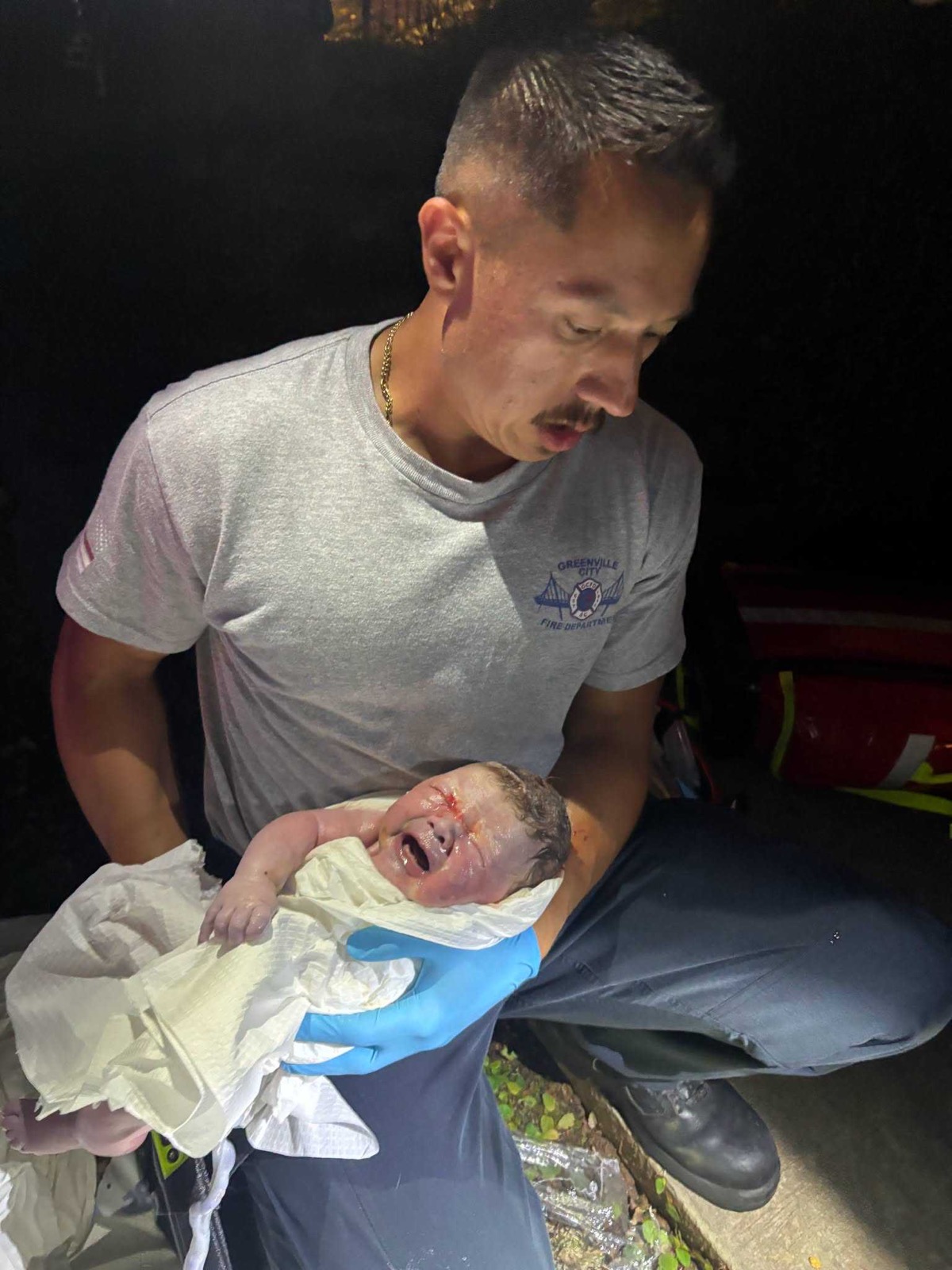 A firefighter in a grey Greenville City Fire Department T-shirt kneels in the dark holding a crying newborn wrapped in a white cloth, one blue-gloved hand supporting the baby