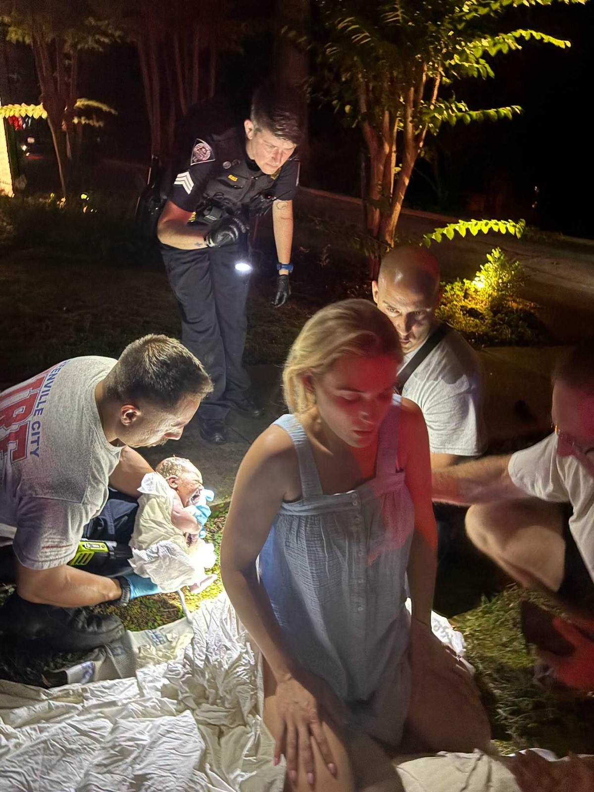 A night-time scene on a lawn: a police officer stands holding a torch, a firefighter kneels holding a newborn wrapped in white, and two more responders crouch beside the mother on sheets spread over the grass